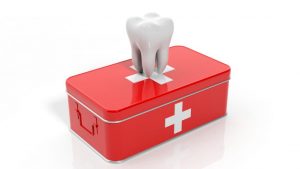A red first aid kit with a white cross on it has a large white tooth model placed on top. The image symbolizes dental care or emergency dental services.