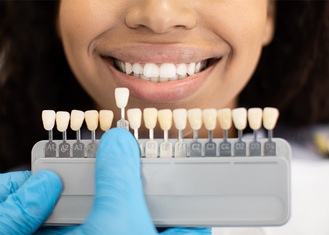 Close-up of a smiling patient with a dental shade guide held near their teeth by a gloved hand, comparing color for cosmetic matching. The guide displays labeled tabs in varying shades, used to select the best match for restorations like crowns or veneers.