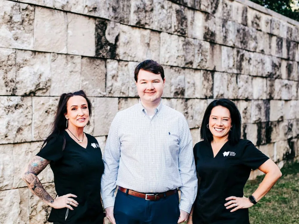 Three individuals stand in front of a stone wall-one wears a light checkered shirt with a brown belt, flanked by two others in black shirts bearing dental care logos. Visible tattoos, a wristwatch, and the tooth-themed branding suggest a casual but professional dental team portrait.