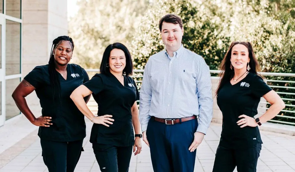 Four people pose outdoors in front of a building with large windows and greenery. Three wear black scrubs with a logo; one wears a light blue checkered shirt and blue pants. Group photo of Stiles Dental Group team